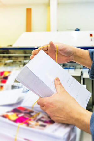 A person putting an elastic band around a stack of printed materials.