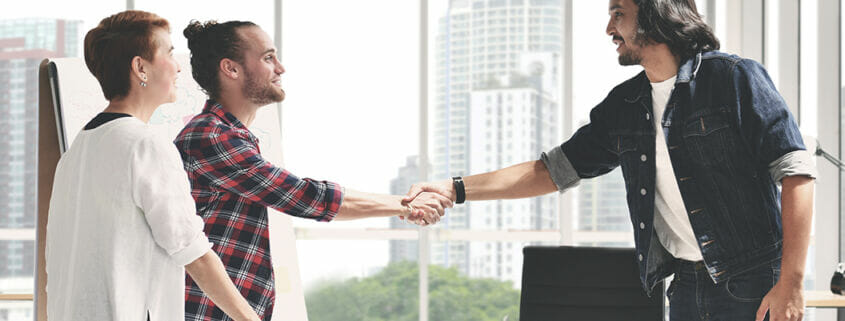 smiling men shake hands in agreement with happy woman standing close by