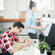 A man sitting at a table looking at a color chart while a woman operates a printing machine.