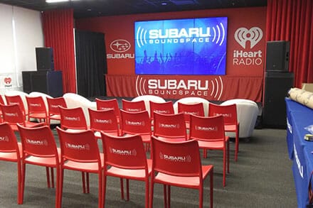 PRI_About-Page-Subaru-iHeart-Radio-Branding Subaru trade show booth with 3 rows of red chairs displaying the Subaru logo.