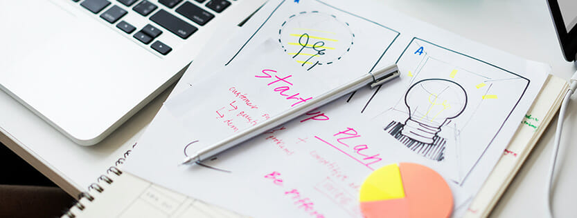 Written ideas for a startup plan on paper next to an apple laptop.